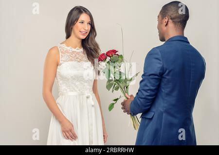 Romantico e affascinante giovane africano che si propone alla sua fidanzata di stringere un mucchio di rose rosse a gambo lungo in mano, con vista sulle spalle Foto Stock