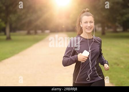 Mezzo Corpo colpo di una bella donna atletica Jogging al parco con le cuffie e sorridente alla fotocamera, con spazio di copia Foto Stock