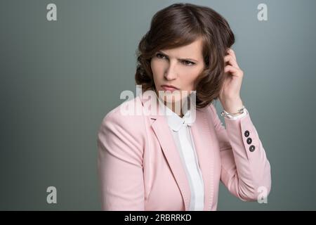 Attraente giovane donna d'affari bruna con un'elegante giacca rosa che le graffiava la testa con un pelo perplesso, su grigio Foto Stock