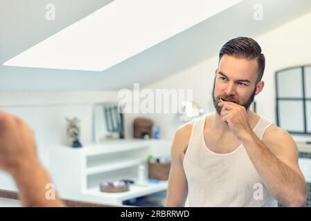 Attraente giovane uomo barbuto spazzolando i suoi denti in bagno a guardare se stesso nello specchio di una igiene dentale concept Foto Stock