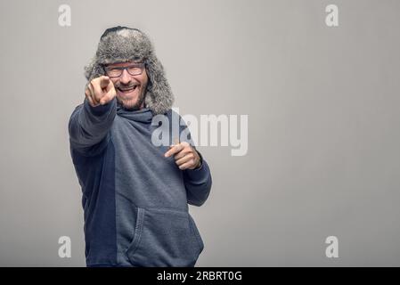 Un uomo gioviale che ride con occhiali e un cappello invernale peloso che punta verso la fotocamera con un'espressione giocosa, in grigio con spazio copioso Foto Stock