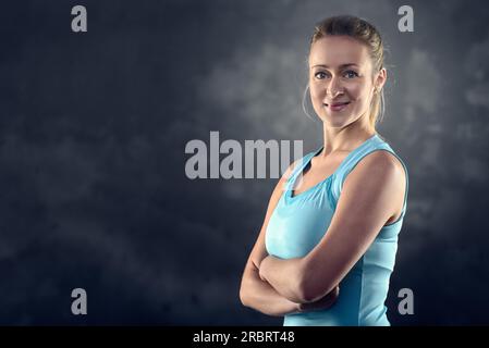 Testa e spalle Ritratto di una donna bionda atletica che indossa una canotta turchese e si trova in piedi con le braccia incrociate in Studio con sfondo grigio Foto Stock