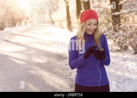 Donna che controlla il suo cellulare per ricevere messaggi di testo con un sorriso felice mentre si gode una sana passeggiata invernale attraverso un parco innevato al sole della mattina presto Foto Stock