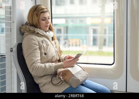 Attraente giovane donna in sella come un passeggero in un treno seduta accanto a una finestra lettura dei messaggi sul suo cellulare con un sorriso, vista di profilo Foto Stock