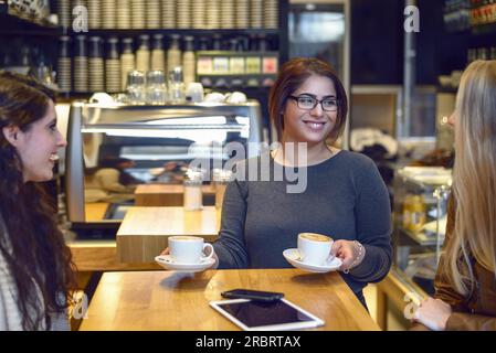 Sorridente cameriera che serve caffè e due giovani donne seduti a un tavolo in un bar o ristorante Foto Stock