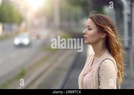 Giovane donna in piedi in attesa in corrispondenza di una stazione ferroviaria sulla deserta open-air platform a fianco di una strada con traffico, vicino la testa e le spalle vista laterale Foto Stock