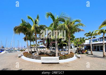 Lanzarote, Isole Canarie, Puerto Calero, ingresso alla principale area dello shopping e dei bar con molte palme di fronte al porticciolo turistico, con un cielo blu intenso Foto Stock