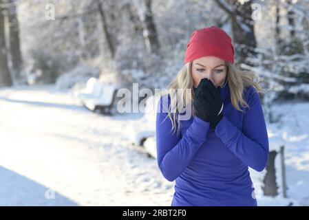 Bella giovane donna bionda che soffia sulle mani dei guanti per tenerli al caldo in un freddo parco invernale sulla neve al sole del mattino Foto Stock