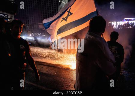 I manifestanti della riforma antigiudiziaria sventolano la bandiera israeliana accanto a un falò che viene estinto dai cannoni ad acqua della polizia sulla strada bloccata di Ayalon durante la manifestazione. Migliaia di israeliani sono scesi in strada e hanno bloccato l'autostrada Ayalon a Tel Aviv scontrandosi con la polizia che ha usato cannoni ad acqua e ufficiali di polizia a cavallo e arresti per disperdere i manifestanti dopo che il capo della polizia di Tel Aviv Amichai Eshed ha annunciato le sue dimissioni dalla forza. Foto Stock