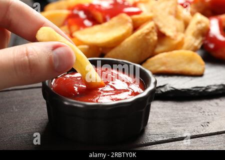 Donna che immerge la frittura francese in una ciotola con il saporito ketchup su un tavolo di legno scuro, primo piano Foto Stock