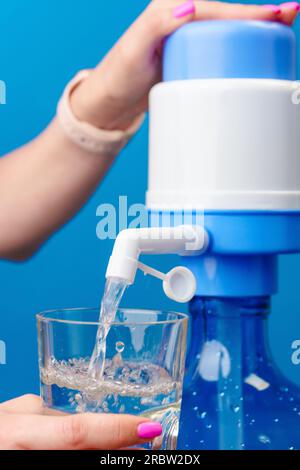 Le mani delle donne versano acqua in un bicchiere dal radiatore dell'acqua su sfondo blu Foto Stock