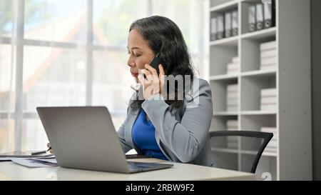 Una donna d'affari asiatica professionale e concentrata è al telefono con il suo partner aziendale alla sua scrivania. Foto Stock