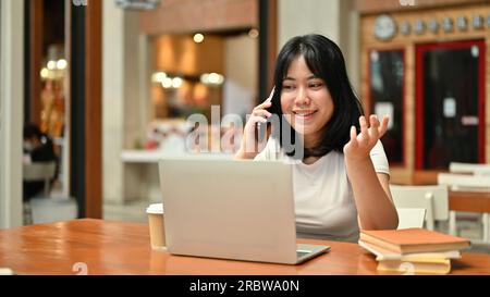 Una giovane e felice freelance asiatica è al telefono con il suo cliente mentre lavora a distanza in un bar. nomade digitale, telelavoro, concetto di stile di vita Foto Stock