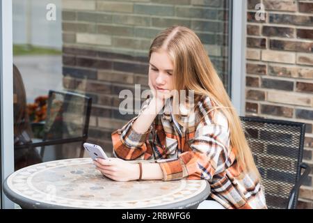 una giovane donna che legge qualcosa al telefono mentre è seduta a un tavolo da una caffetteria all'aperto Foto Stock