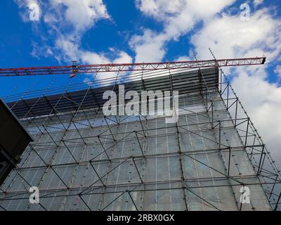 Gru e ponteggi intorno a un alto edificio industriale in un cantiere edile. Vista dall'angolo basso, cielo blu, niente persone. Foto Stock