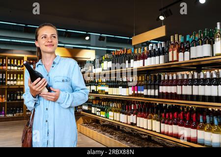 Giovane donna bruna adulta che sceglie vino in enoteca e sorride. Foto Stock