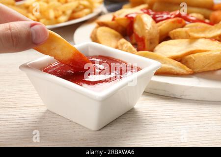 Donna che immerge deliziose patate al forno in un recipiente con ketchup al tavolo di legno, primo piano Foto Stock