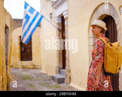 Giovane donna asiatica in abito rosso etnico cammina e guarda le strette stradine della città vecchia. Turismo, vacanza e scoperta concetto, viaggiatore femminile in visita sud Europa, isola di Rodi, Grecia. Foto Stock