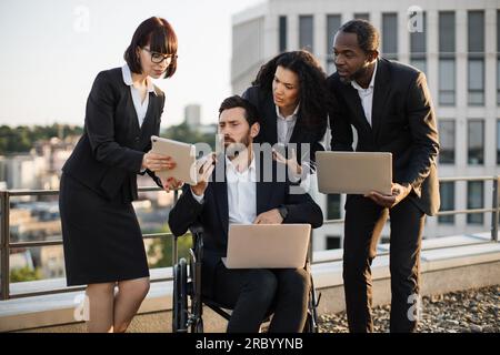 Imprenditori multirazziali in outfit aziendali che discutono di strategia aziendale fuori ufficio. Partner aziendali che si riuniscono intorno a un uomo disabile in Whecha Foto Stock