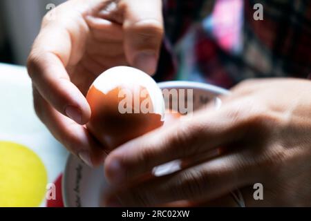 Vista ravvicinata delle mani che tengono e sbucciano un uovo di guscio di uovo duro bollito in una ciotola sul tavolo all'interno di una casa a colazione UK KATHY DEWITT Foto Stock