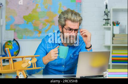 Insegnante maschio serio che beve caffè o tè in classe. Insegnante o professore universitario sul posto di lavoro. Insegnante di scuola in occhiali seduti alla scrivania Foto Stock