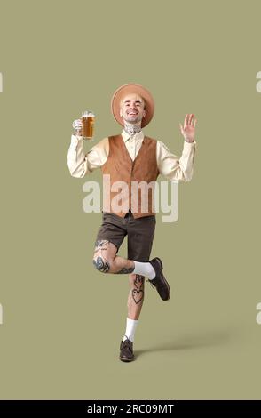 Giovane uomo tatuato in abiti tradizionali tedeschi con birra che balla su sfondo verde Foto Stock