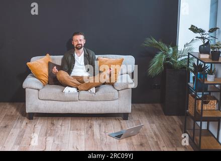 Uomo seduto a casa sul divano, adottando la posa del loto asana e abbracciando lo stato di relax. Con un'espressione serena sul volto, si immerge nella pratica della meditazione o della consapevolezza. Foto di alta qualità Foto Stock