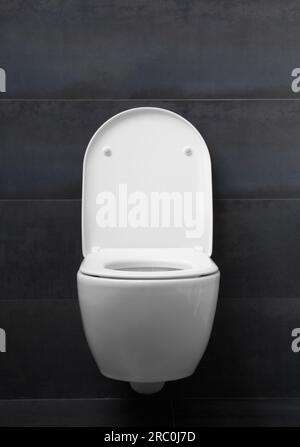 Un moderno WC bianco montato a parete con sedile abbassato, coperchio rialzato e pulsante cromato lucido a filo contro una parete nera. WC in bagno Foto Stock