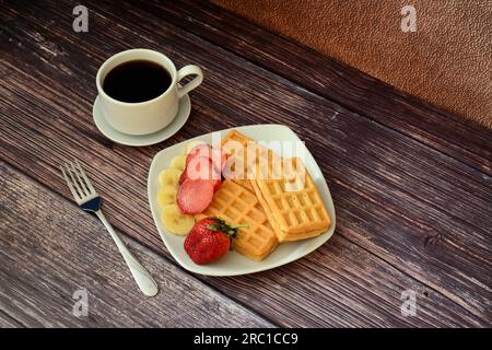 Una tazza di caffè nero su un piattino, una forchetta e un piatto con waffle freschi viennesi con fragole e banane su un tavolo di legno. Vista dall'alto, base piatta. Foto Stock