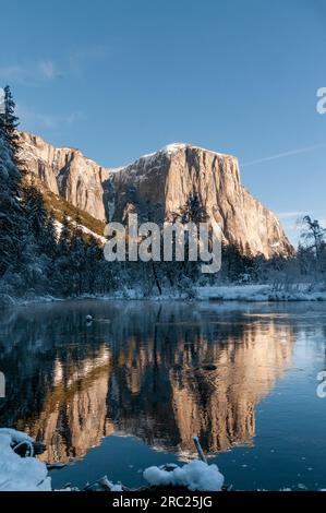 La superficie rocciosa delle montagne della Sierra Nevada, che riflette la luce del sole del mattino presto, si riflette nel fiume Merced, in un paesaggio innevato di inizio inverno nel parco nazionale di Yosemite. Foto Stock