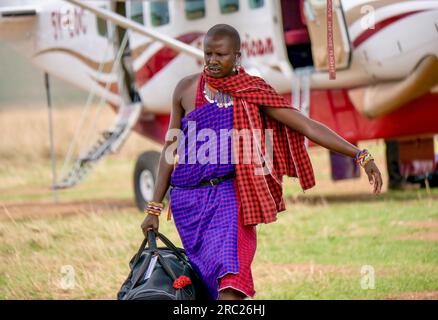 Maasai Mara, Kenya - 25 settembre 2013. Un membro del personale del campo safari Masai porta una borsa passeggeri da un aereo charter su una pista erbosa. Foto Stock