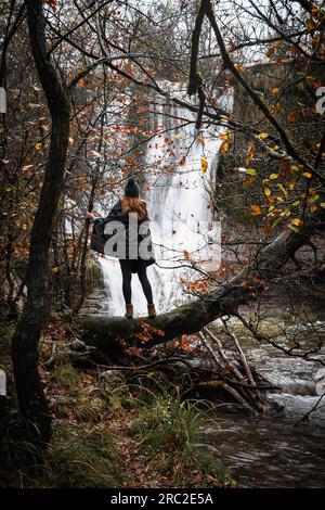paesaggio di una cascata in autunno con una giovane donna che la osserva tra gli alberi rossastri Foto Stock