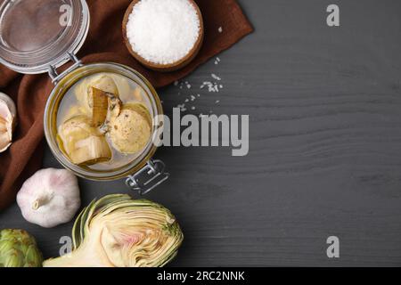 Vasetto di carciofi deliziosi sottaceto in olio d'oliva e ingredienti su tavola di legno grigio scuro, piatto. Spazio per il testo Foto Stock