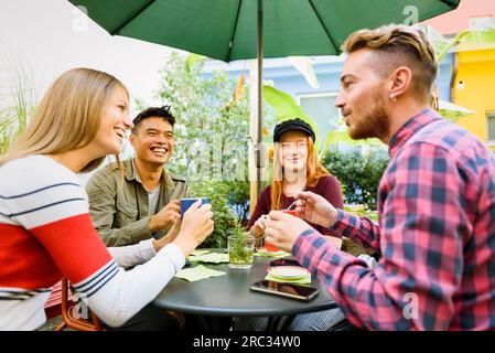 Uomini e donne multirazziali in abiti informali sorridono e parlano tra loro mentre si siedono al tavolo durante la pausa caffè nella caffetteria all'aperto Foto Stock