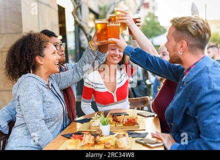 Gruppo di amici multirazziali che si trovano all'esterno dell'edificio con spuntini su un tavolo di legno con telefoni cellulari e che guardano in alto mentre bevono un bicchiere di birra Foto Stock