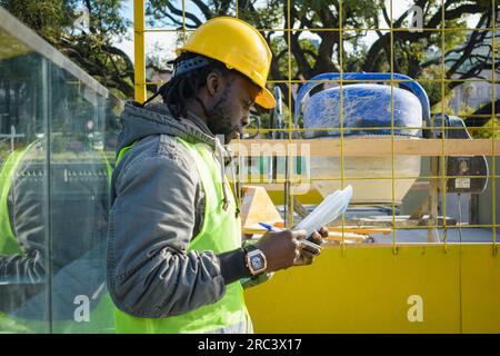 Giovane ingegnere di etnia africana che indossa giubbotto e casco di sicurezza, in piedi all'esterno del cantiere leggendo le istruzioni e le attività per Foto Stock