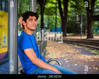 Tre quarti di foto di un bellissimo giovane uomo in ambientazione urbana che aspetta il tram alla fermata dell'autobus, indossando una polo blu Foto Stock