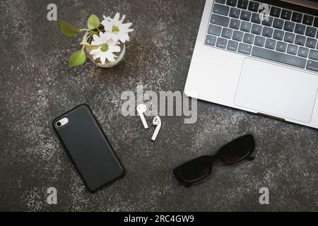 Vista orizzontale dell'area di lavoro. Dotato di computer portatile, telefono, cuffie, occhiali da sole e pianta su scrivania scura. Foto Stock