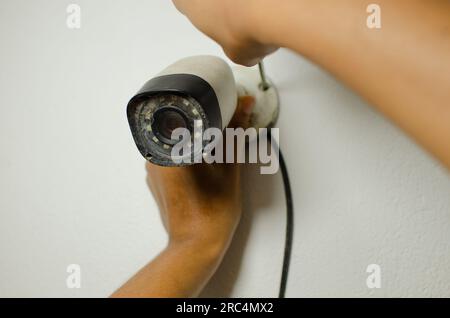 Primo piano di mani professionali che eseguono la manutenzione su una telecamera di sicurezza bianca, rafforzando il concetto di protezione e sorveglianza. Foto Stock