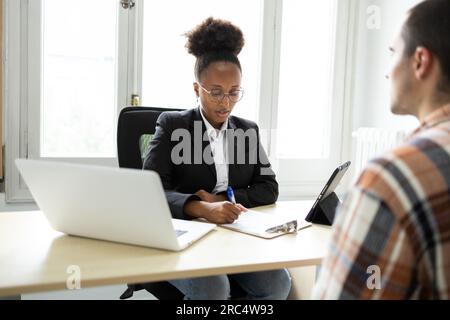 Giovane avvocato afro-americana in occhiali da vista che guarda le note sugli appunti mentre si siede al tavolo con un computer portatile, un tablet e un interattico Foto Stock