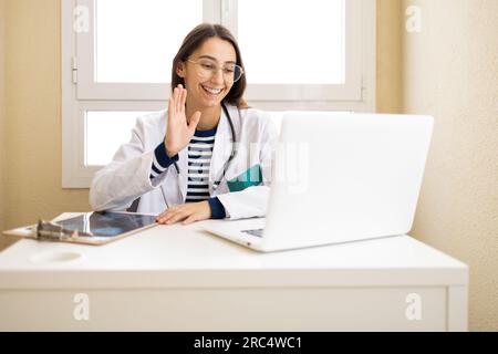 Giovane medico positivo in mantello bianco che salta la mano al paziente durante una videochiamata sul computer portatile in ufficio Foto Stock