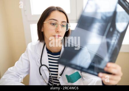 Giovane medico positivo in mantello bianco e occhiali che leggono raggi X mentre si siede al tavolo in ufficio Foto Stock