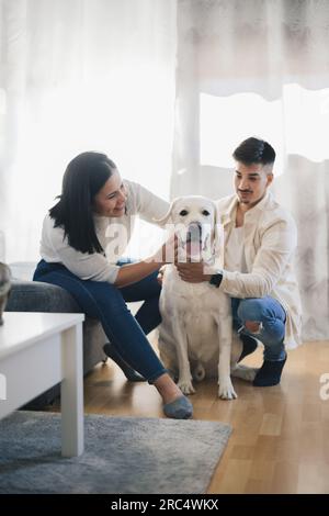 Coppia felice in abiti informali che accarezza e posa con Labrador Retriever in salotto a casa su sfondo sfocato Foto Stock