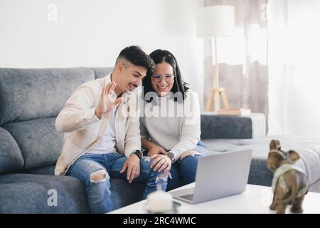 Felice giovane coppia in abiti informali seduti sul divano in salotto guardando lo schermo mentre si effettua una videochiamata con il computer portatile Foto Stock