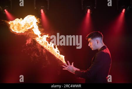 Vista laterale del prestidigitatore maschile in costume nero che mostra un trucco magico con fiamme che si esibiscono sul palco oscuro del circo Foto Stock