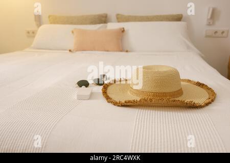 Cappello e occhiali da sole sul letto dell'hotel con cuscini bianchi e beige e coperta durante le vacanze estive nelle Isole Baleari Foto Stock