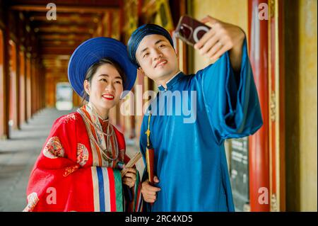 Felice coppia asiatica in abiti e copricapi tradizionali sorridente e scattante selfie mentre si trova in piedi su uno sfondo sfocato del passaggio dell'edificio a Hue Vietn Foto Stock
