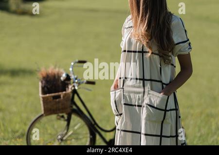 Coltiva una giovane donna senza volto irriconoscibile in abito bianco con lunghi capelli in piedi vicino alla bicicletta con cesto pieno di fiori su prato verde Foto Stock