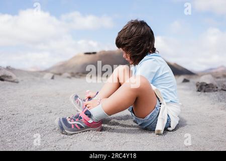 Vista laterale di un anonimo preadolescente in abiti casual che guardano in basso seduto su un terreno asciutto e legano i lacci delle scarpe alla luce del sole contro il cielo blu nuvoloso Foto Stock