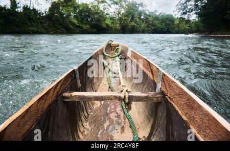 Vecchia barca di legno con corda legata da prua a tavola di legno che galleggia nelle acque blu del fiume dell'Ecuador vicino a alberi verdi contro il cielo blu nuvoloso Foto Stock